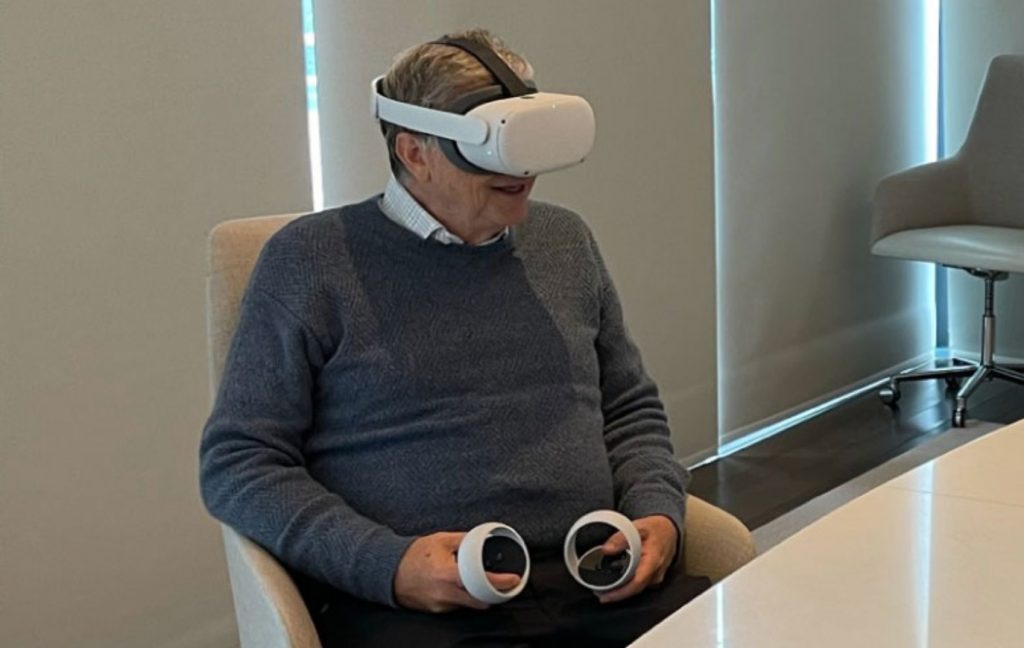 Bill Gates sentado em uma cadeira, segurando controles e um óculos de realidade virtual na cabeça