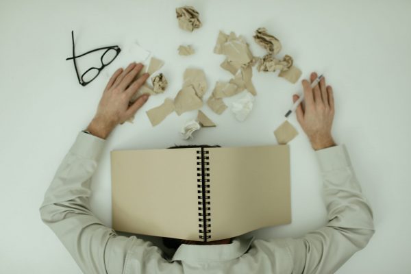Imagem demonstra o burnout, o esgotamento emocional, representado por uma pessoa debruçada sobre uma superfície lisa, com um caderno aberto cobrindo a sua cabeça, papéis espalhados ao redor, além de óculos