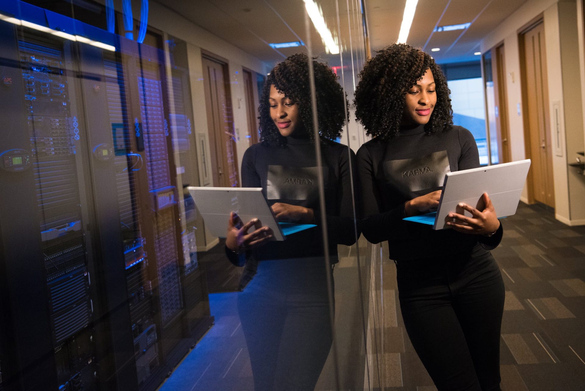 Na imagem, uma mulher negra segurando um notebook encostada em uma parede de vidro representa carreira em tecnologia para mulheres