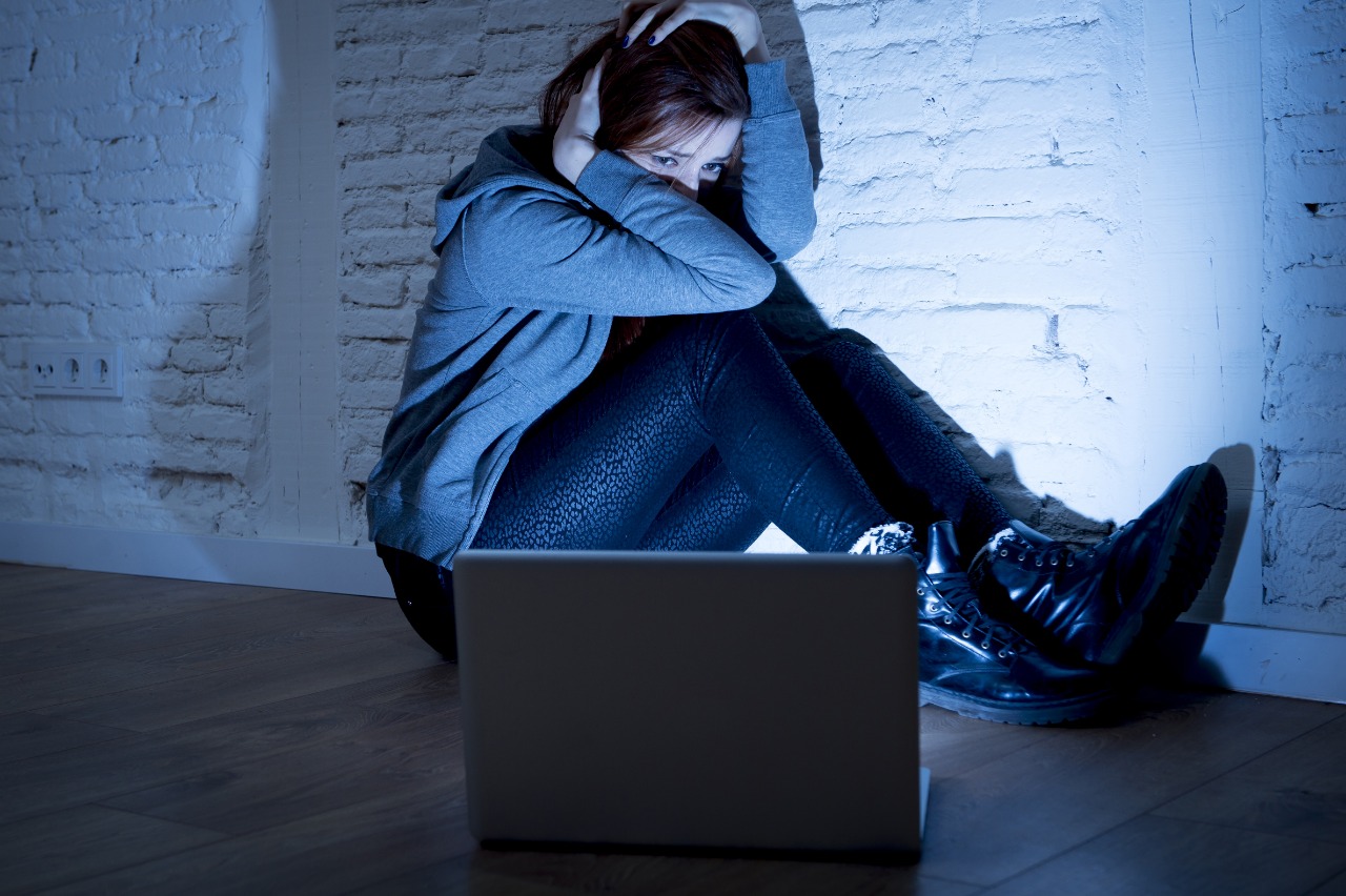Imagem mostra uma mulher sentada em uma sala escura, com o rosto escondido entre os braços, olhando para a tela de um computador como se tivesse sido vítima de revenge porn