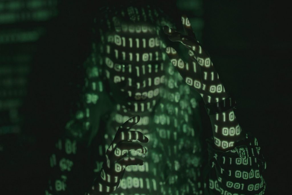Imagem mostra a silhueta de uma pessoa com uma projeção de códigos de computador em sua face; a imagem é esverdeada em fundo escuro
