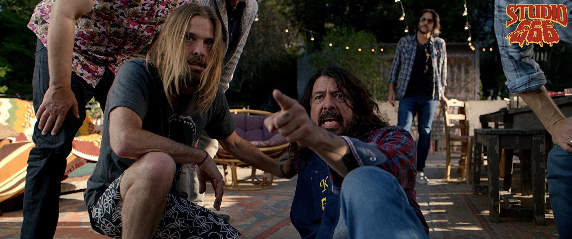 Cena do filme Studio 666, do Foo Fighters; cena mostra o vocalista Dave Grohl sentado no chão, apontando o dedo indicador para a direção da câmera, ao lado aparece o baterista Taylor Hawkins agachado