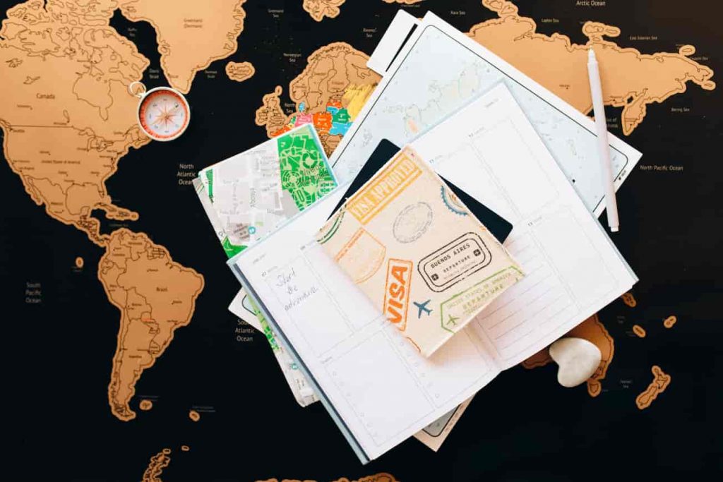 Ao fundo é possível ver um mapa mundial colocado em uma superfície plana, por cima está uma bússola, uma agenda e um passaporte empilhados
