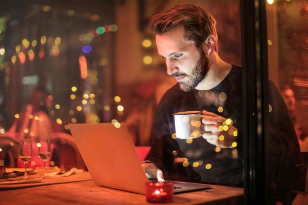 Homem nômade digital sentado em um café segurando uma caneca com a mão esquerda e digitando algo em um notebook com a mão direita