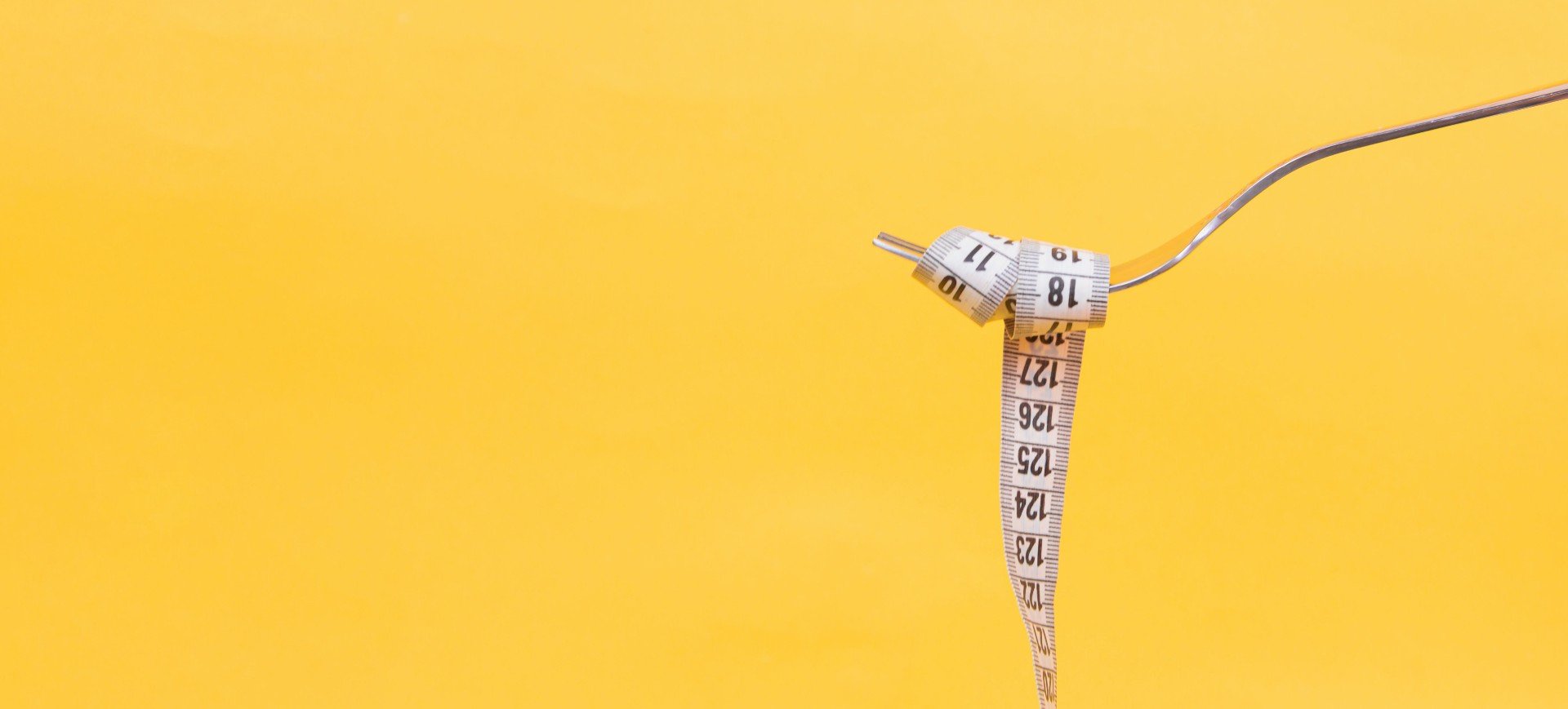 Uma fita métrica enrolada em um garfo, como um macarrão, em uma imagem de fundo amarelo; imagem ilustra transtorno alimentar