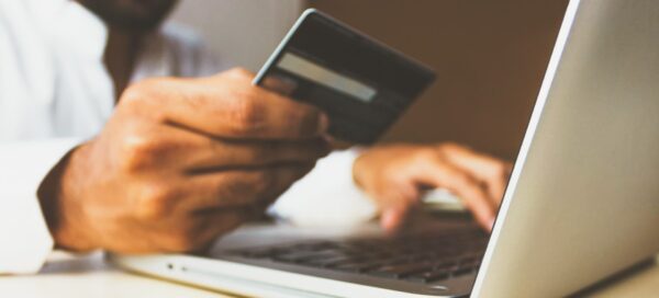 Imagem mostra uma homem segurando um cartão de crédito com a mão direita e digitando algo em um notebook com a esquerda, como se estivesse finalizando uma compra on-line