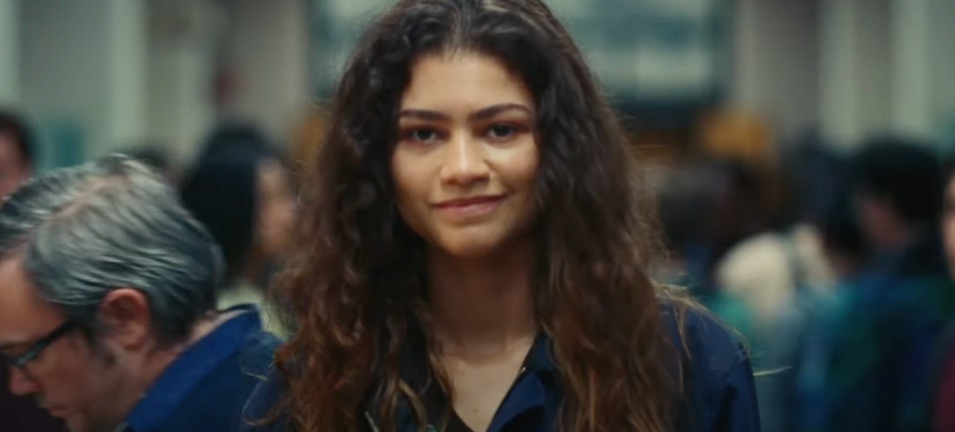 Uma cena da temporada 2 de Euphoria: na frente, Rue Bennet, personagem de Zendaya, aparece com um sorriso tímido; ao fundo há uma porção de pessoas aleatórias passando