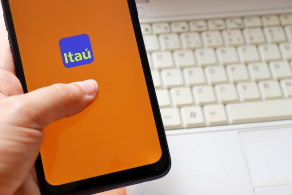 Na imagem, uma mão segura um smartphone com o logotipo do banco Itaú na tela; ao fundo há um teclado de notebook