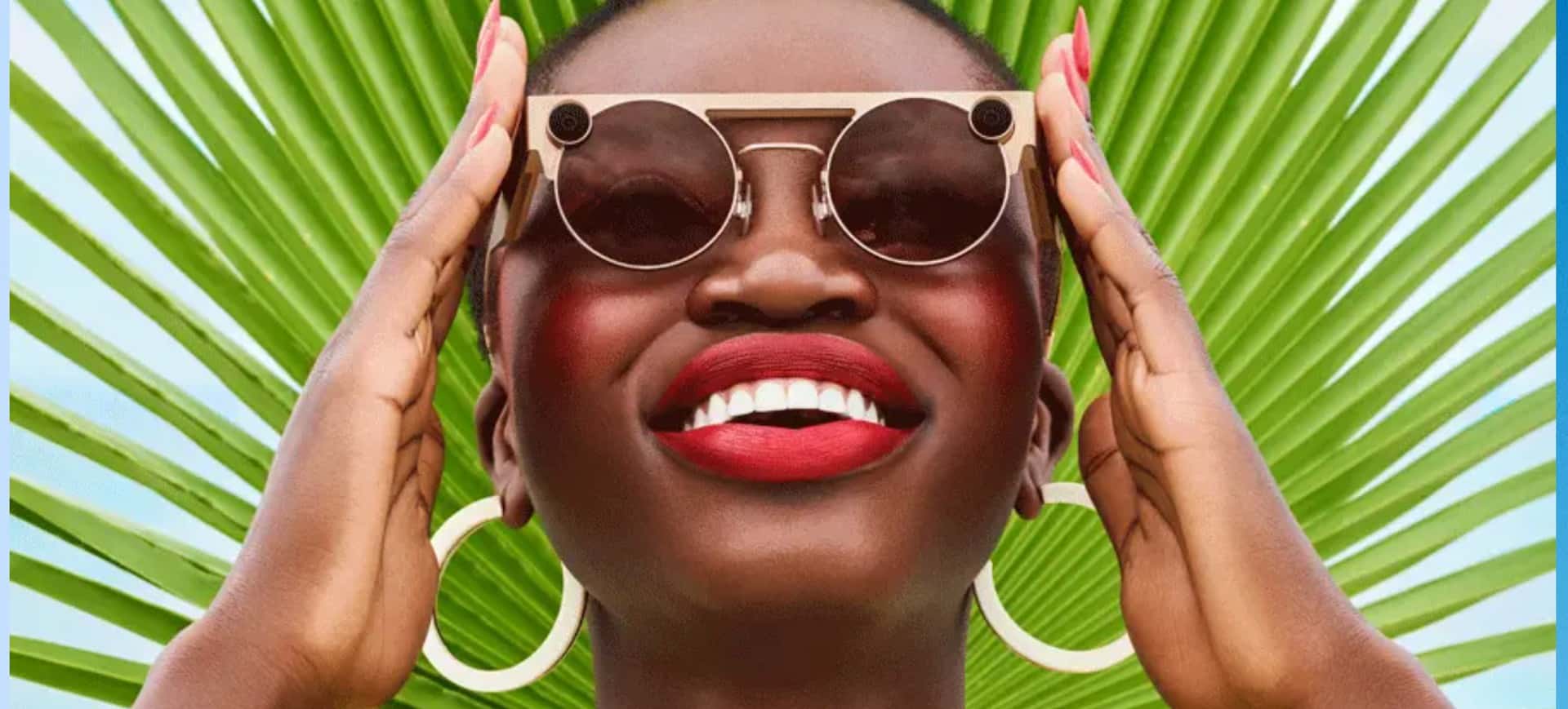 Rosto de uma mulher negra, que utiliza um batom vermelho, veste brincos de argola e os óculos de realidade virtual da Snap, o Spectacles; as mãos da mulher aparecem ao lado dos óculos, segurando a haste e o fundo da imagem possui dois tons de verde intercalados