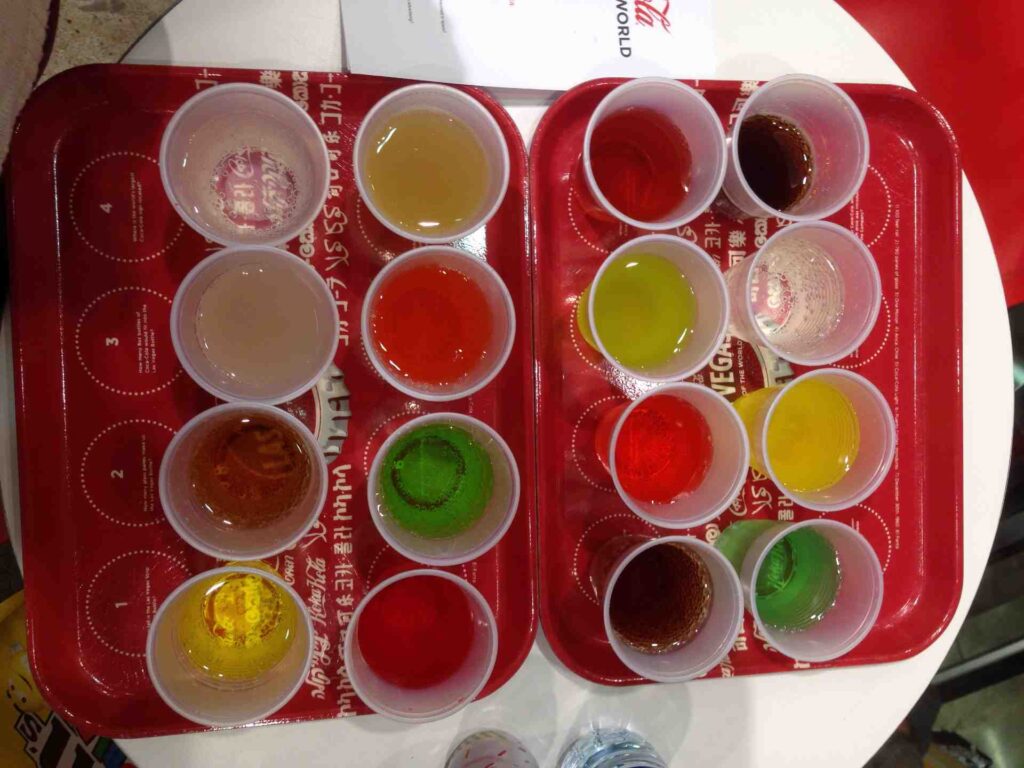 Em uma bandeja vermelha já 16 copos, todos com líquidos coloridos que são os sabores das Coca-Colas pelo mundo, incluindo a brasileira Juat