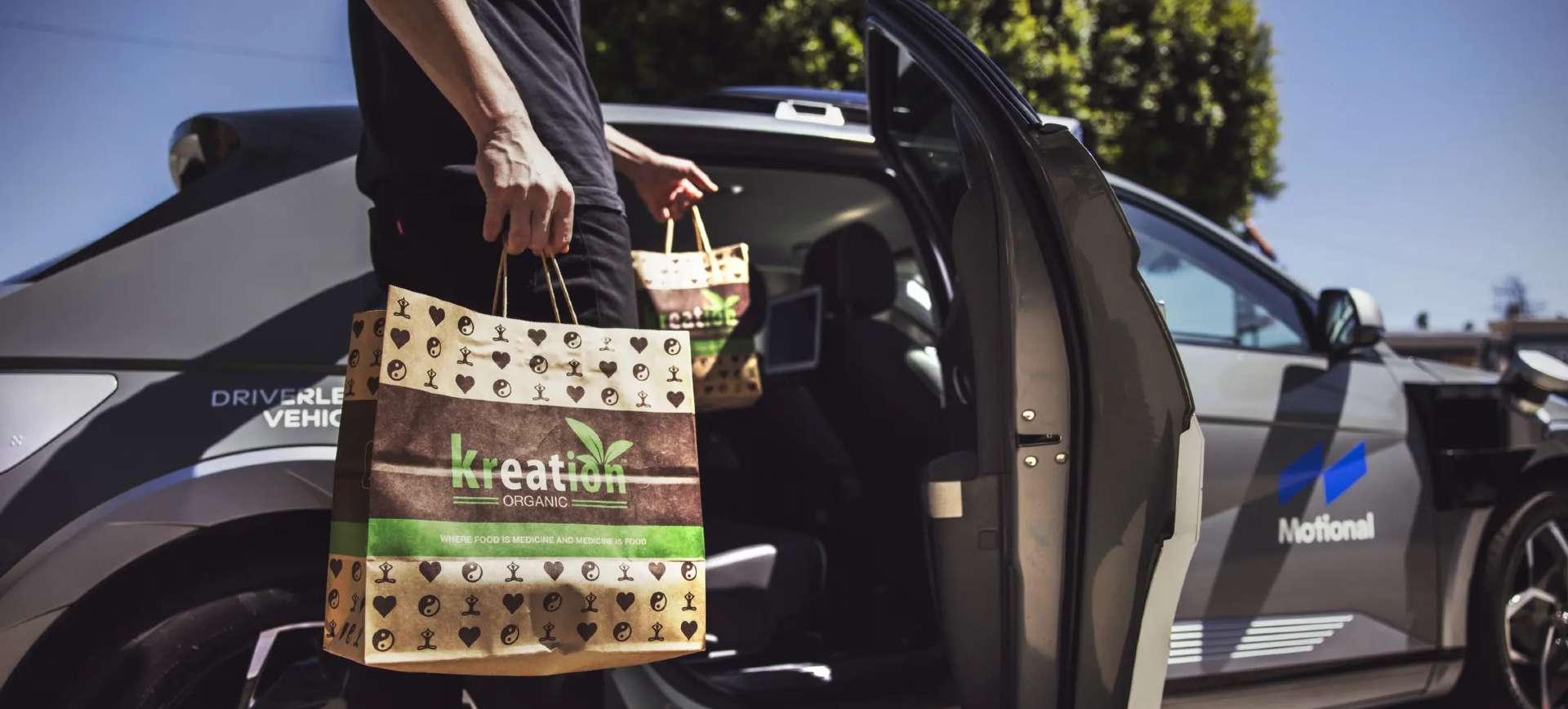 Na imagem aparece o torço do corpo de um homem, segurando sacolas para colocá-las dentro de um carro autônomo da Motional, que iniciou testes de delivery autônomo em parceria com a Uber Eats