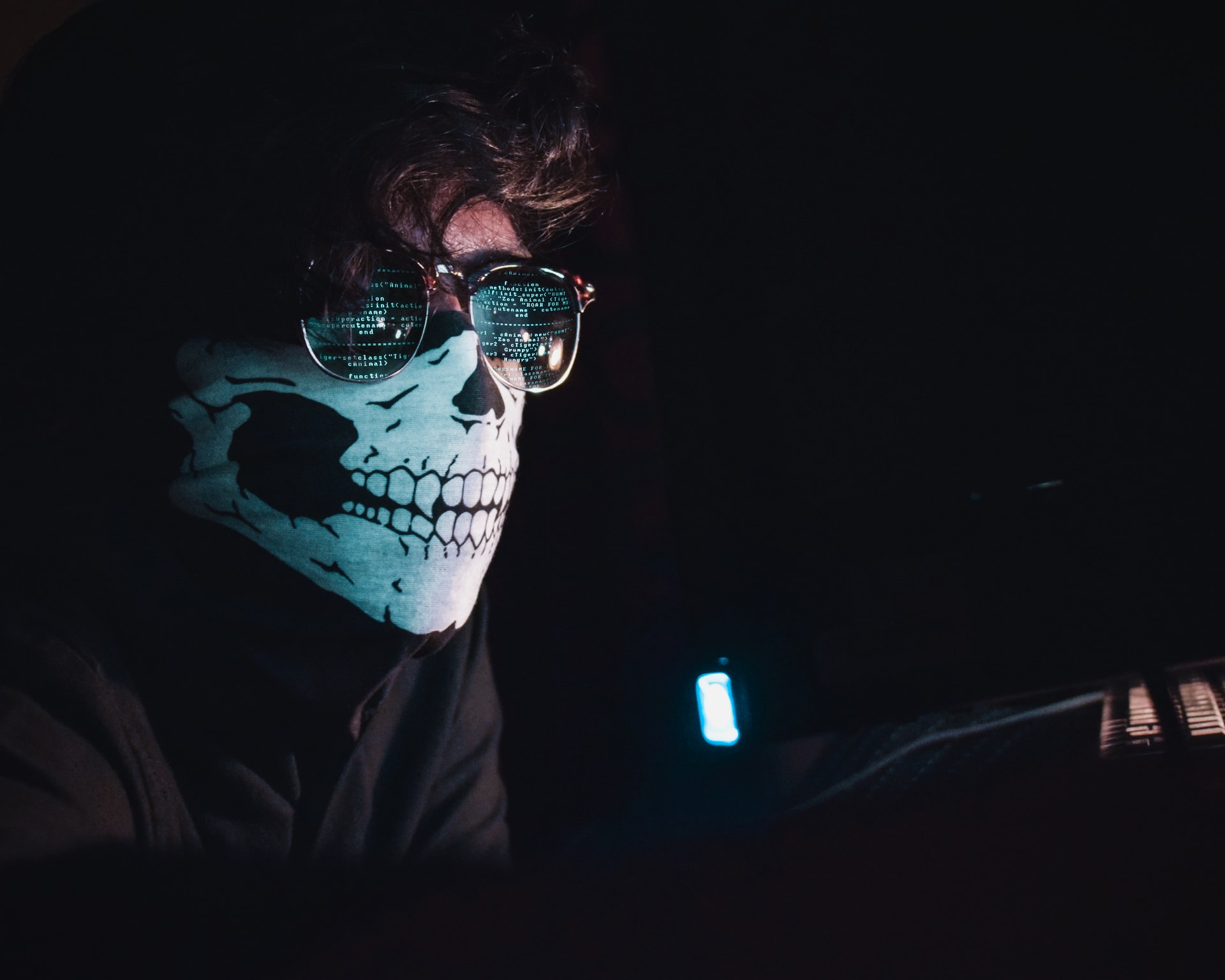 Imagem mostra um homem de máscara e óculos escuros à frente de um computador. Nas lentes dos óculos, está refletida uma tela que mostra várias linhas de códigos de programação