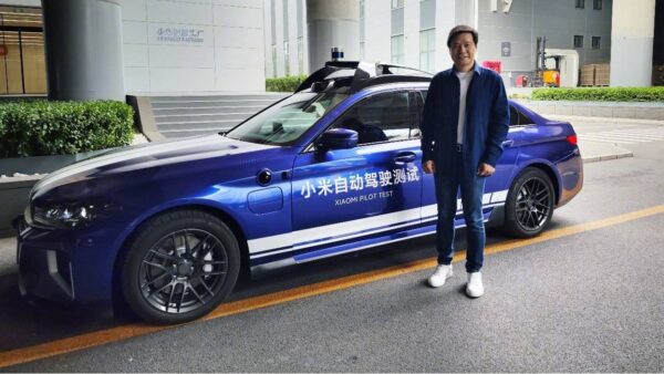 Foto mostra o CEO da Xiaomi, Lei Jun, posando ao lado de um protótipo do Pilot, o carro que a empresa vem desenvolvendo para o setor de automóveis elétricos. O veículo tem a cor azul, com adesivos sinalizando sua natureza de protótipo. Lei Jun veste jaqueta e jeans azuis com tênis brancos e aparece sorrindo para a câmera.