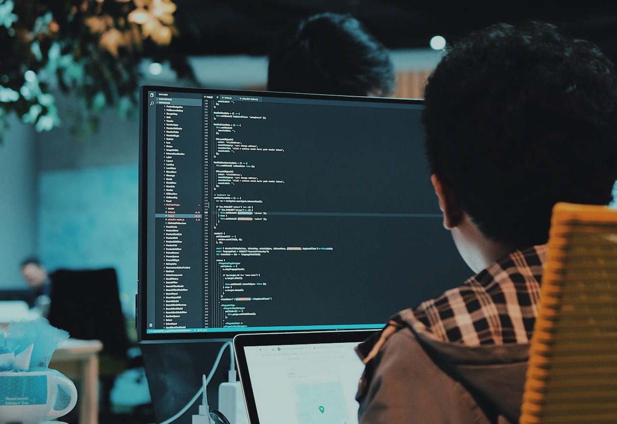 Imagem mostra as costas de um menino programador que está de frente para um computador, na tela é possível ver os códigos de programação que ele está desenvolvendo
