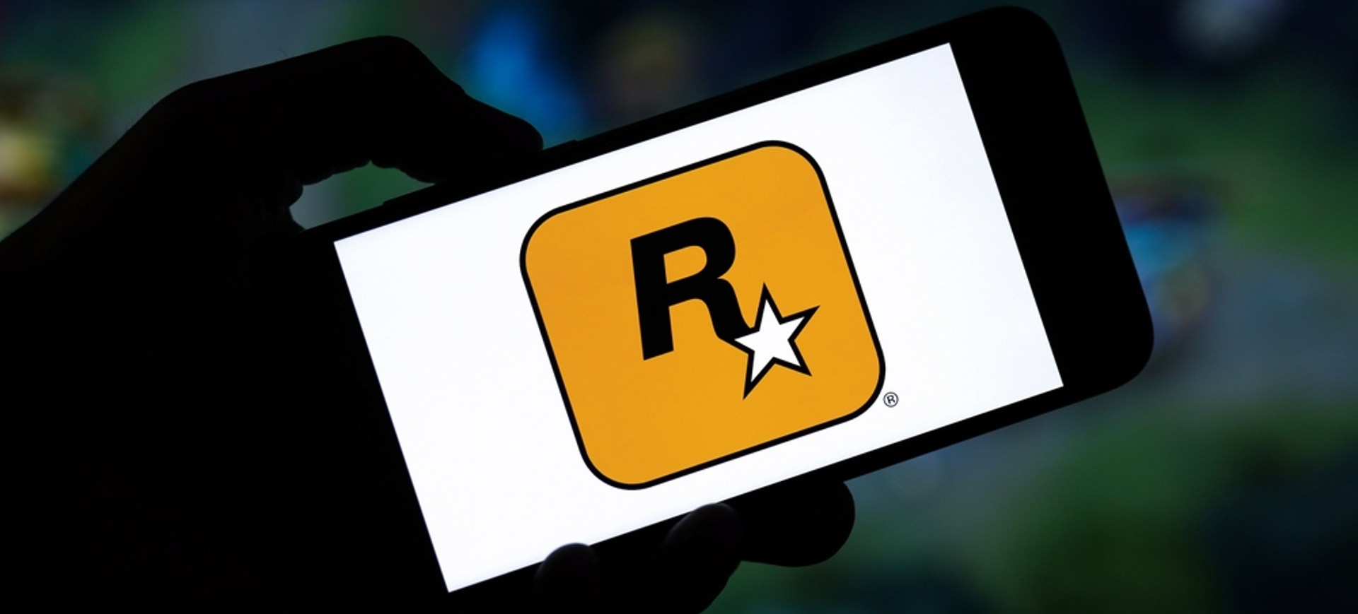 A sombra de uma mão segura um smartphone, o qual exibe em sua tela o logo da desenvolvedora Rockstar Games