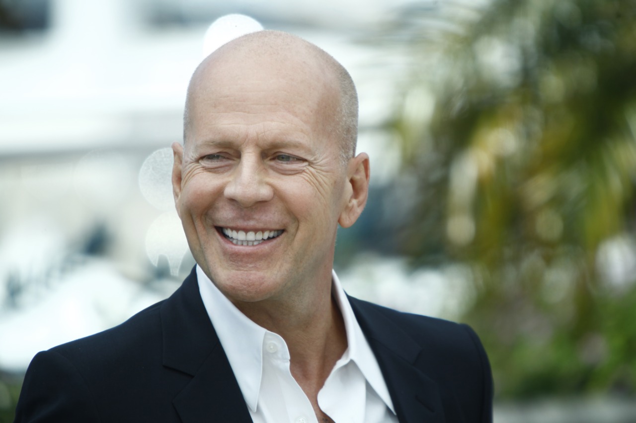 Imagem do ator Bruce Willis sorrindo para a câmera, vestindo um paletó preto, com camisa branca e sem gravata