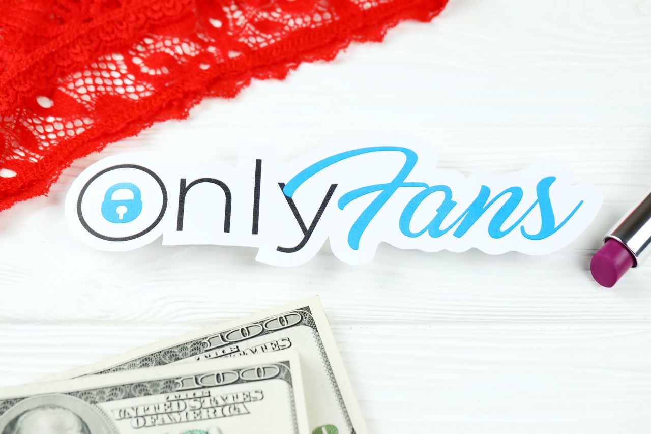 Imagem mostra adesivo com logotipo do OnlyFans, com notas de dólares embaixo da tela, um batom vermelho à sua direita e uma lingeria também vermelha por cima