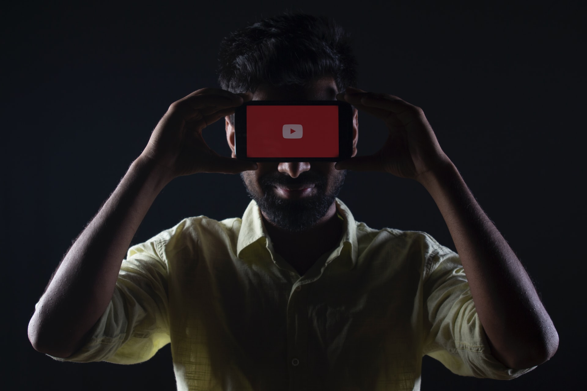 Foto mostra um homem segurando um smartphone na altura dos olhos: o celular tampa os olhos do homem da visão da câmera e, em sua tela, está o símbolo do YouTube