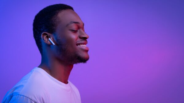 Imagem de um homem negro à frente de um fundo lilás iluminado. Ele sorri de olhos fechados enquanto ouve músicas com o fone de ouvido