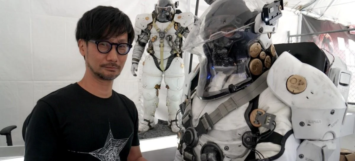 Imagem mostra Hideo Kojima ao lado de uma estátua em tamanho humano do robô símbolo do estúdio Kojima PRoductions