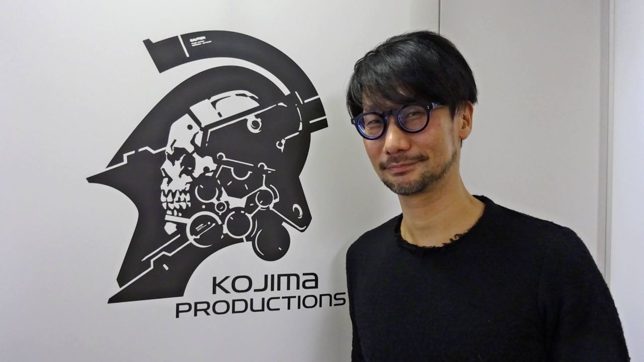 Imagem mostra Hideo Kojima ao lado de uma parede com o símbolo do estúdio Kojima Productions