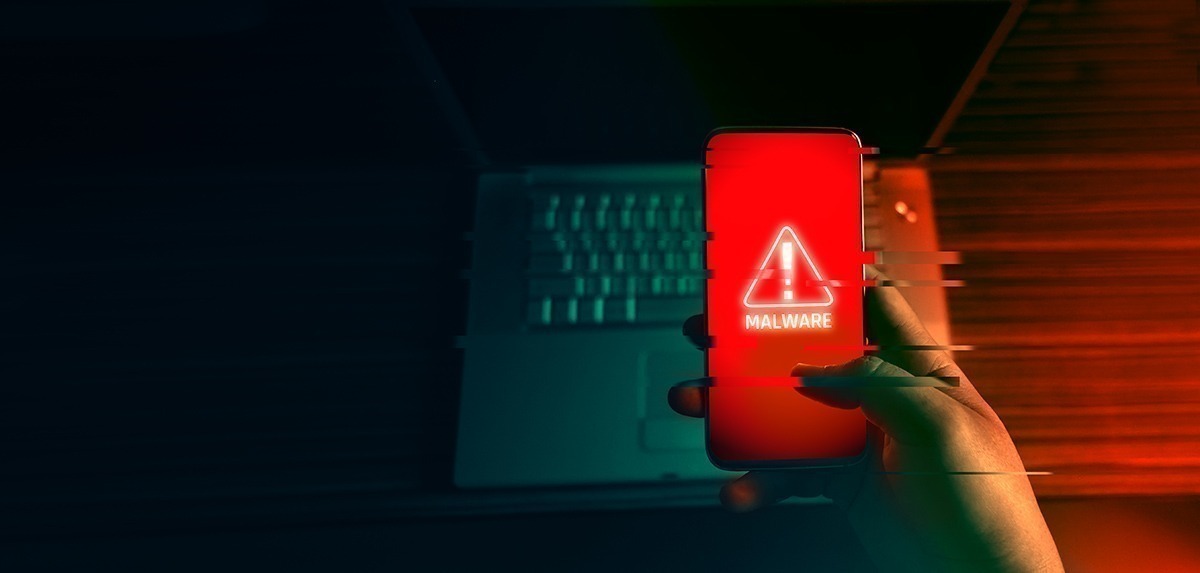 Imagem mostra um celular com alerta vermelho e um notebook ao fundo, ambos infectados por malware