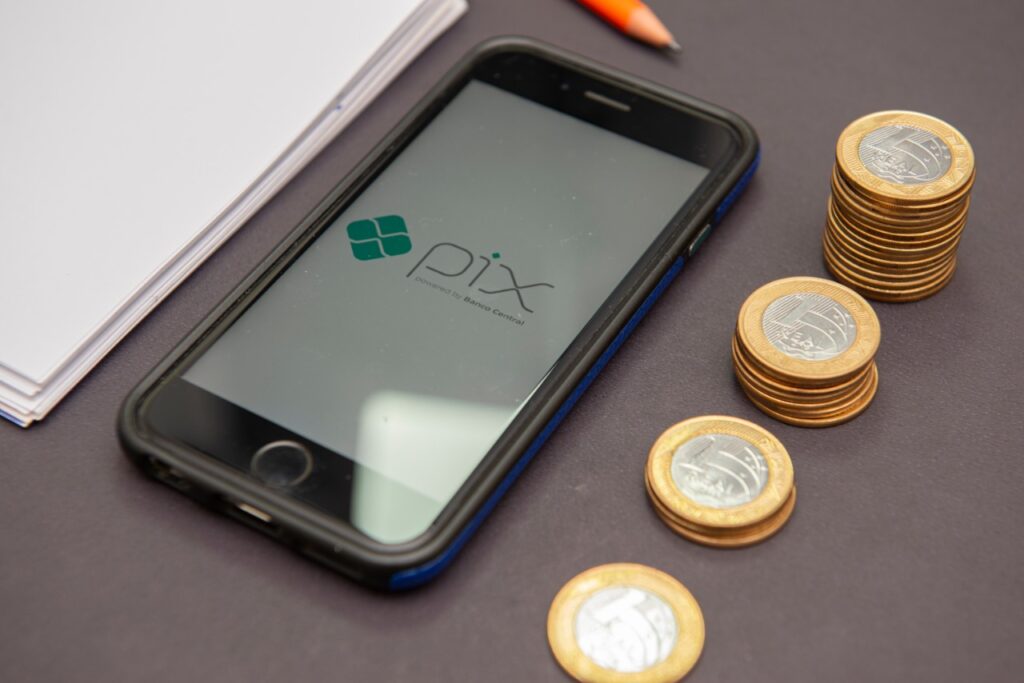 Imagem mostra um celular com logotipo do Pix e várias cédulas e moedas do Real em volta