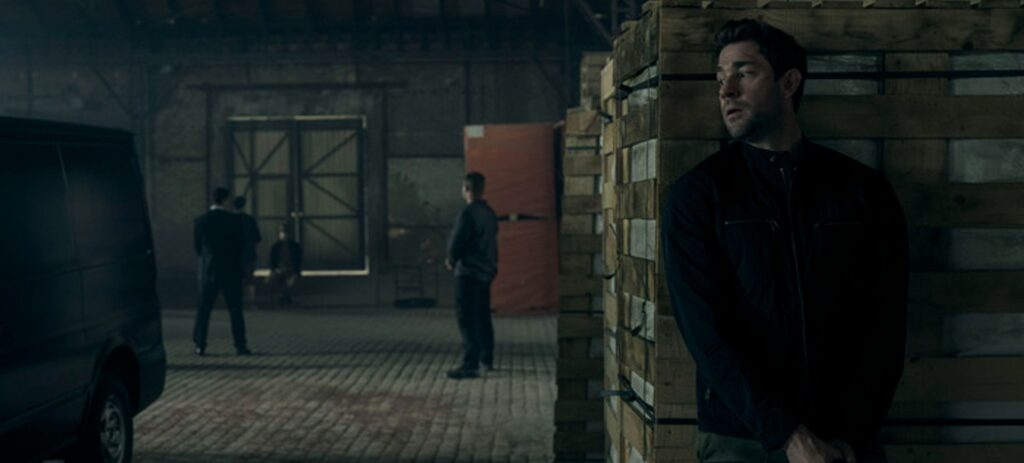 Cena da série Jack Ryan de Tom Clancy, do Prime Video - na foto, o ator John Krasinski que dá vida ao protagonista escondido atrás de caixas em um galpão, portando uma arma