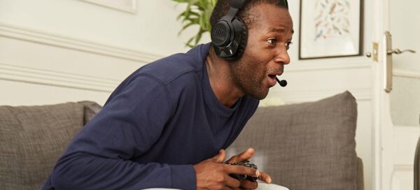 Imagem mostra um jovem adulto homem sentado em um sofá creme, vestindo uma camiseta azul marinho de mangas longas e o fone de ouvido JBL Quantum 360X e segurando um controle de videogame
