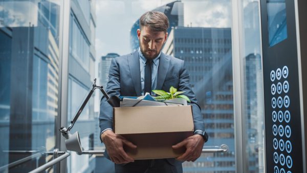 Imagem mostra um homem de paletó carregando uma caixa com pertences, simbolizando demissões de empresas