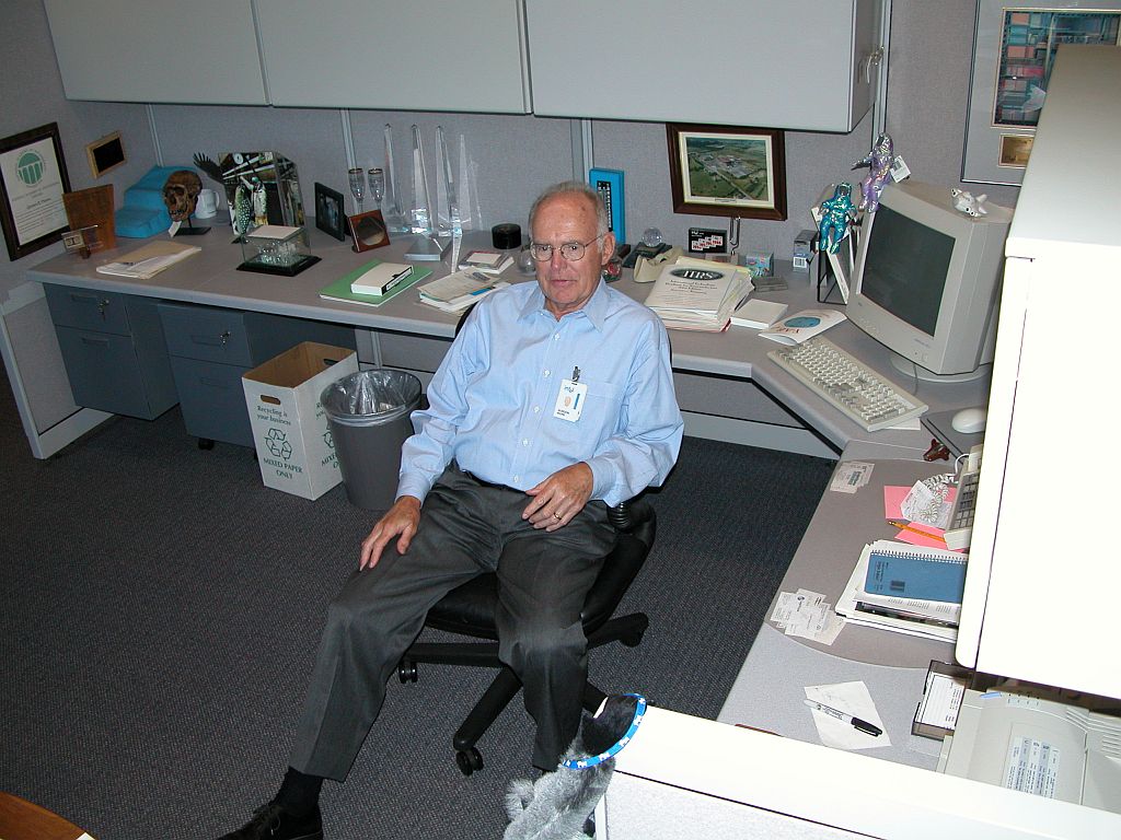 Gordon Moore, pai da lei de Moore e um dos fundadores da Intel, vestido com calça social e camisa azul claro, sentado em uma cadeira em seu escritório na empresa