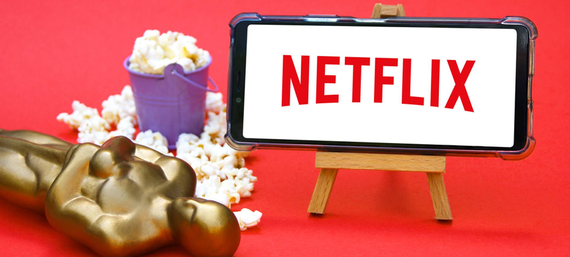Em um fundo vermelho, a imagem mostra uma estatueta do Oscar deitada no chão, ao lado de um pote de pipoca e um cavalete segurando um smartphone, cuja tela mostra o logotipo da Netflix