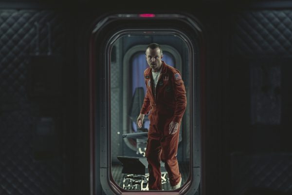 Na imagem, o ator Aaron Paul veste um macacão espacial laranja e aparece em uma abertura de porta; cena da temporada 6 de Black Mirror