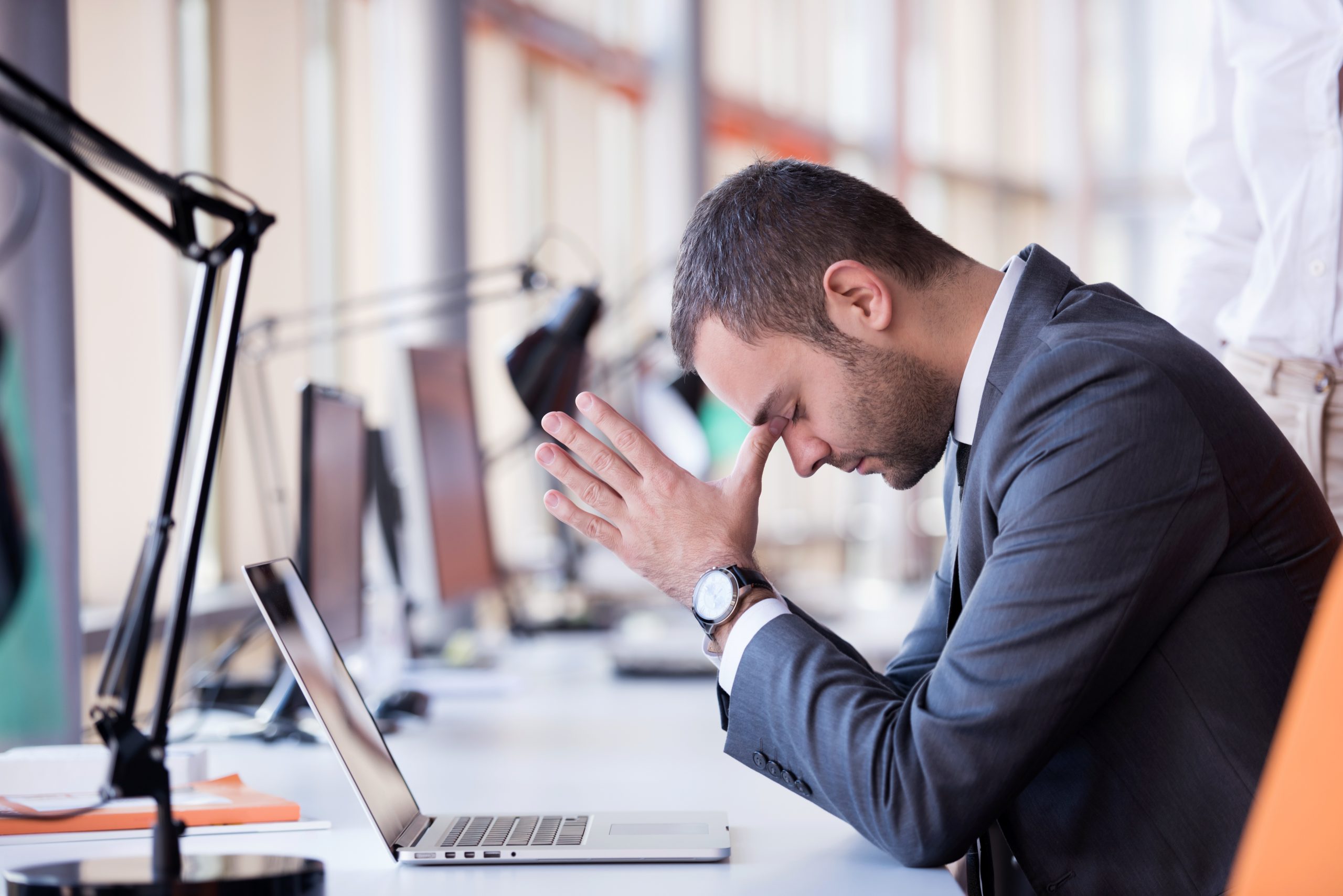 Imagem mostra homem de paletó e relógio com as mãos sobre o rosto, simbolizando stress no ambiente de trabalho