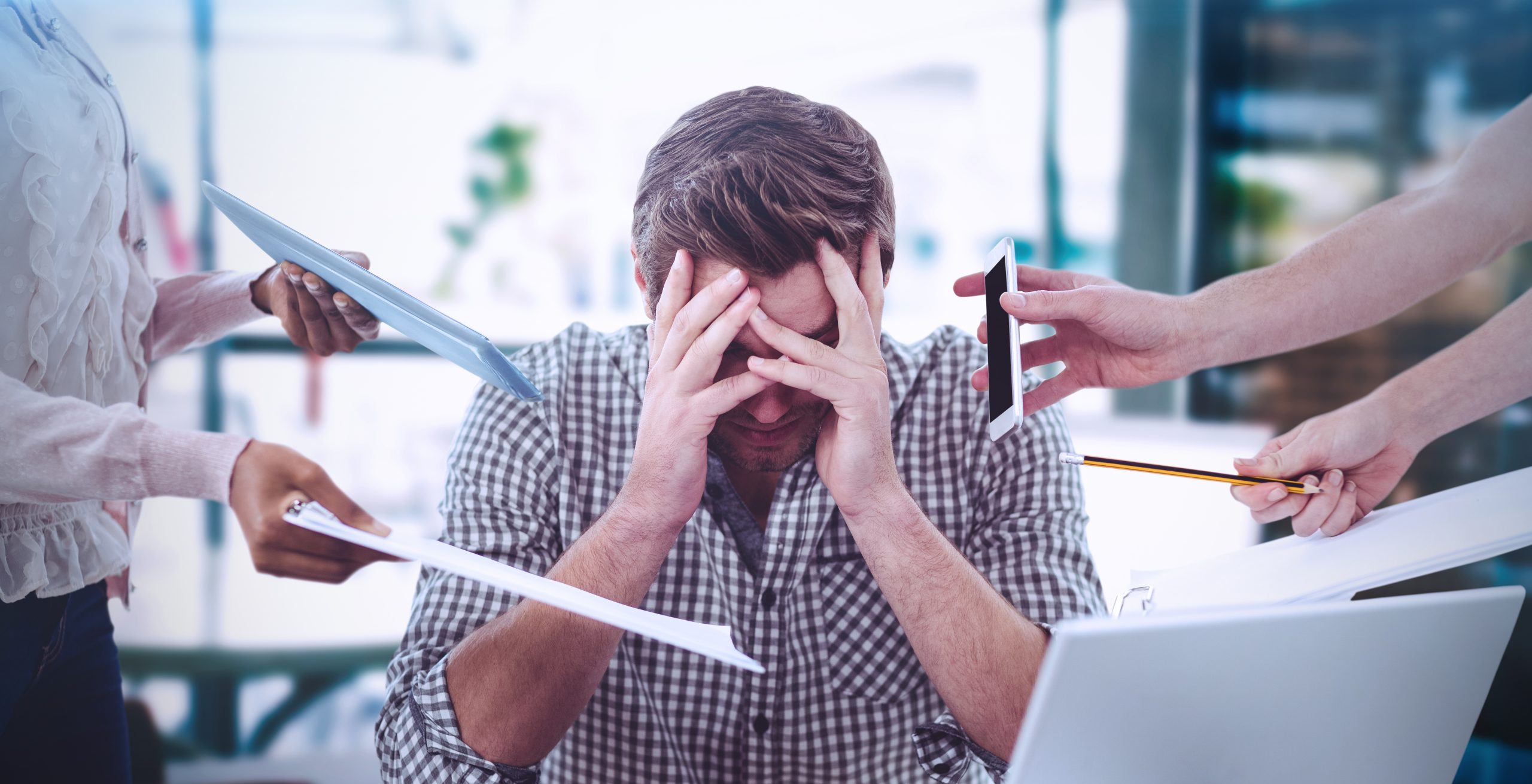 Imagem mostra homem de camisa com as mãos sobre o rosto, simbolizando stress no ambiente de trabalho. Ele está sendo constantemente interrompido por outras pessoas