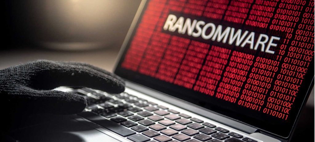 Imagem de uma mão com luvas pretas digitando algo no teclado de um notebook, na tela do dispositivo aparece a palavra ransomware