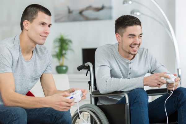 Imagem mostra dois homens, sendo um cadeirante, se divertindo com um videogame, remetendo à acessibilidade nos jogos