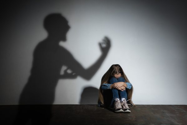 Imagem mostra uma criança ajoelhada no chão, escondendo o rosto com medo, enquanto a sombra de um adulto está ao lado dela, ameaçando-a