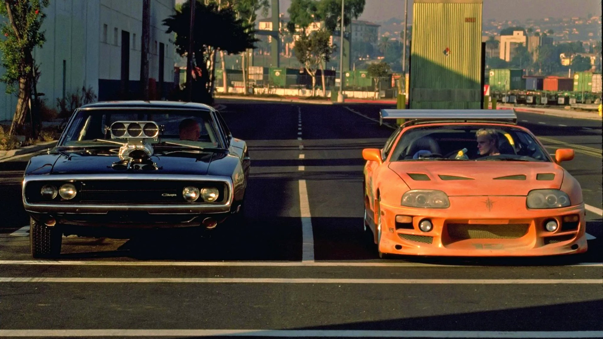 Imagem mostra cena do primeiro filme da saga Velozes e Furiosos, com dois carros, um preto americano e um laranja japonês, prestes a começar uma corrida de rua