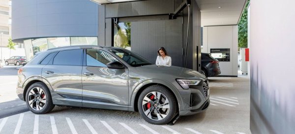 Foto mostra por dentro da estação de carregamento de carros elétricos da Audi na Áustria; na imagem há uma mulher caucasiana, de cabelos longos e lisos, vestida com uma camisa branca e de pé, ao lado de um carro cinza da marca, realizando a recarga da bateria do veículo