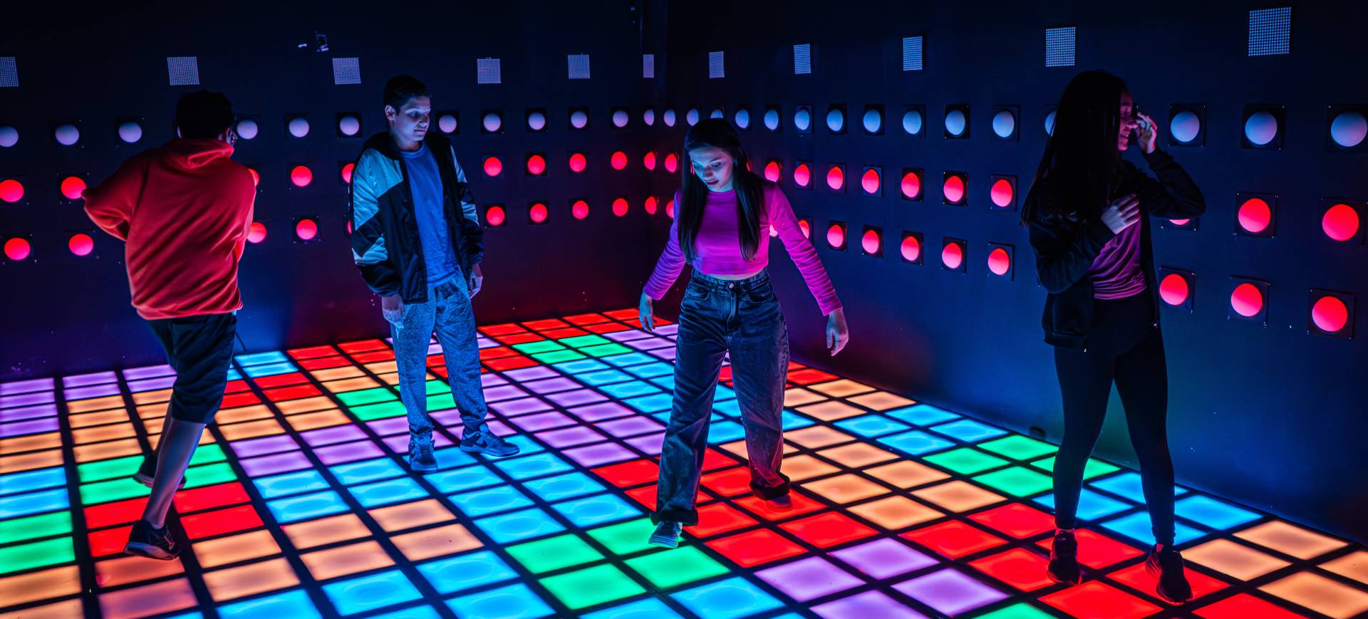 Imagem de divulgação da Arcade Haus, um parque tecnológico de imersão, equipado com botões e luzes de LED coloridos, que aparecem no chão e paredes da foto e com os quais quatro jovens estão interagindo
