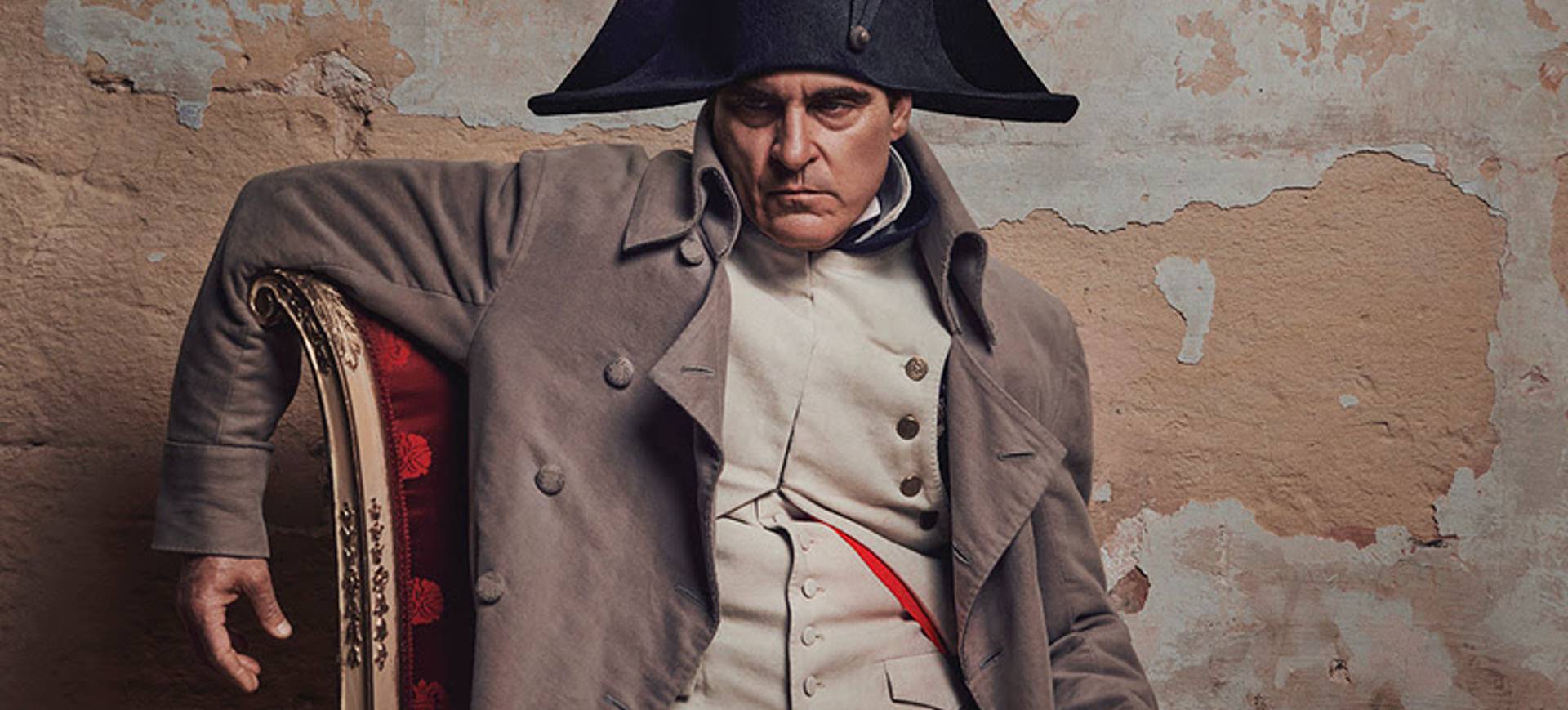 Pôster do longa Napoleão, que aparece o ator Joaquin Phoenix vestido como o imperador com seu icônico chapéu, sentado em uma cadeira