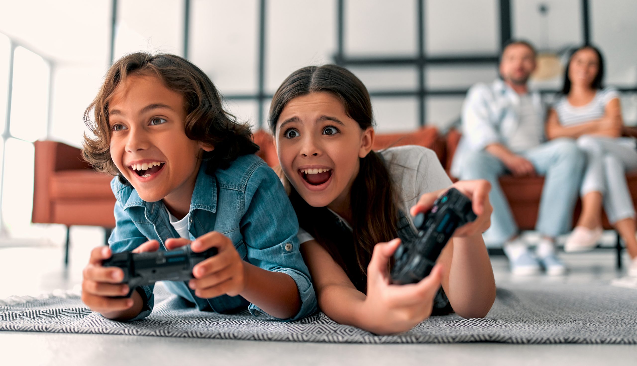 Imagem mostra duas crianças jogando videogame com os pais observando-as ao fundo