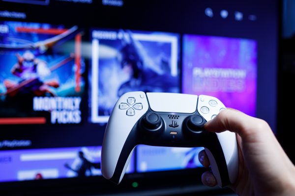 Imagem mostra o controle DualSense do PlayStation 5 segurado por um jogador e uma smart TV ao fundo