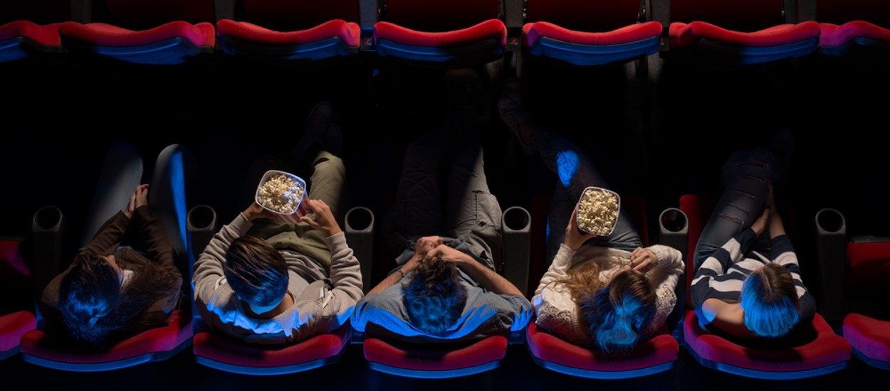 Jovens sentados no cinema, assistindo a um filme e comendo pipoca; imagem de vista superior ilustra a Semana do Cinema