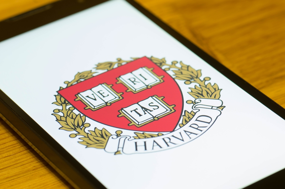 Na imagem, um celular cuja tela exibe o logotipo da Universidade de Harvard; foto ilustra o curso gratuito fornecido pela Fundação Estudar, com conteúdo baseado no CC50 de Harvard, introdutório e com foco em programação