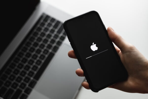 Mão segura um iPhone que, por sua vez, exibe o logo Apple na tela, mostrando uma atualização do sistema. Ao fundo, o teclado de um MacBook. Imagem ilustra atualização de segurança em sistemas da Apple