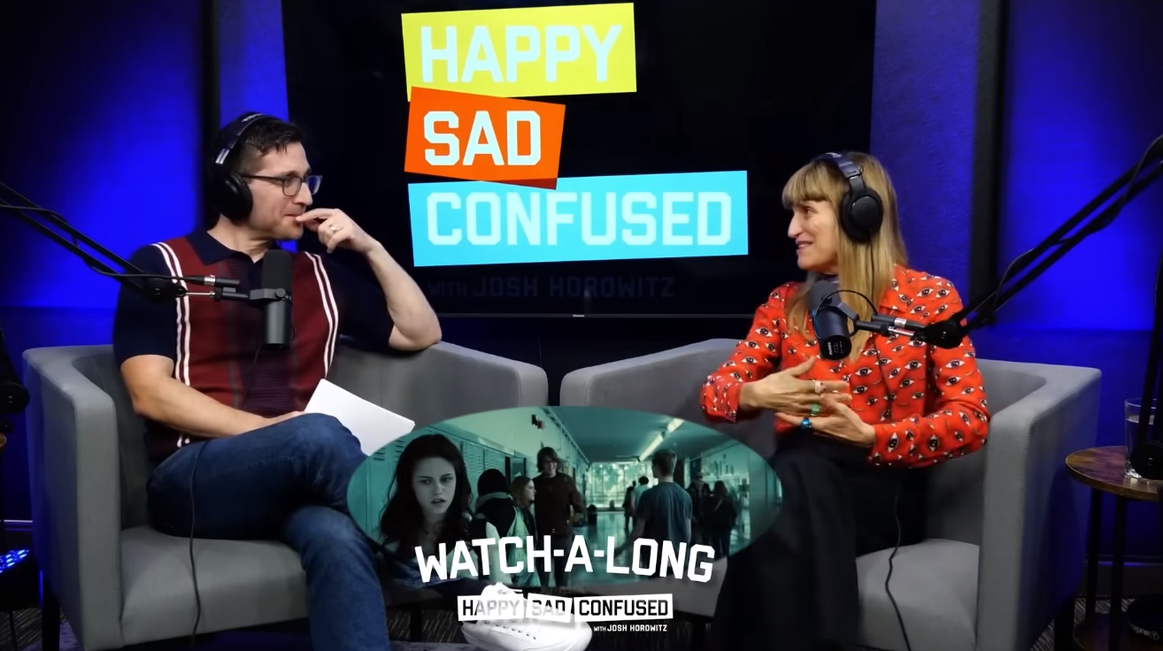 Cena com Josh Horowitz entrevistando Catherine Hardwicke, diretora dos filmes da saga Crepúsculo, durante o podcast Happy Sad Confused; ambos estão sentados em poltronas, com headsets e microfones, em uma sala de estúdio de áudio