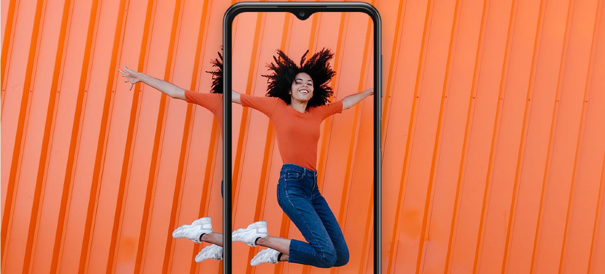 Imagem conceito com a cor laranja de fundo, à frente mostra o smartphone Samsung A23. Na tela, a imagem de uma mulher dando um leve salto; ela está vestida com calça jeans e blusa de manga três quartos laranja, e tênis branco