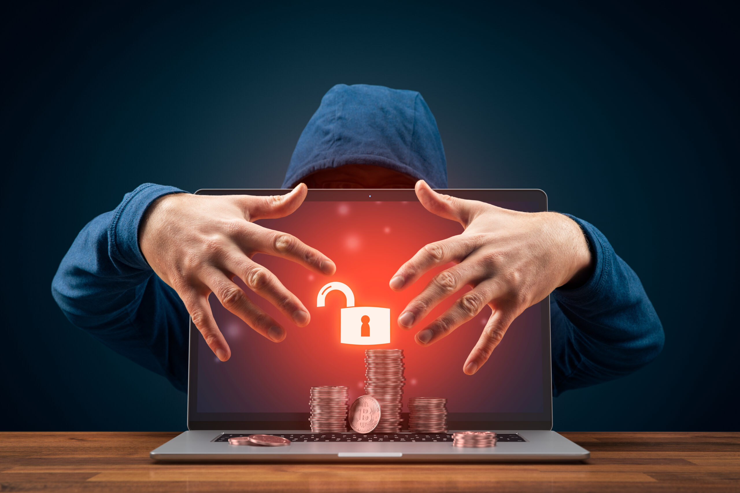 Imagem de um hacker com as mãos em cima de um laptop com moedas à frente